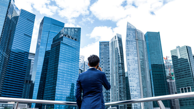 A person in a blue suit stands on a balcony, looking at a skyline of modern skyscrapers under a partly cloudy sky.