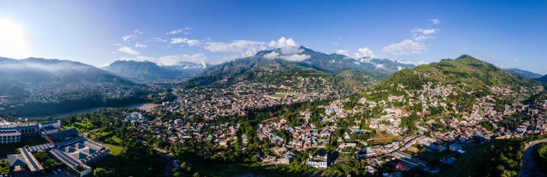 Aerial view of the sprawling city nestled in a lush valley between majestic green mountains under a clear blue sky with a winding river Muzaffarabad, Azad Kashmir, Pakistan.