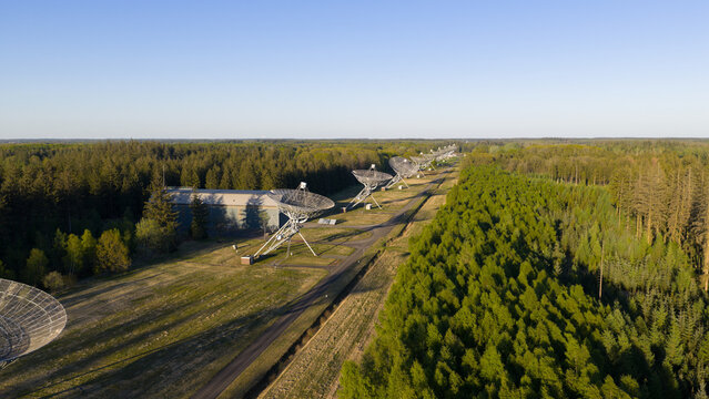 Aerial view of the Westerbork Synthesis Radio Telescope dishes lined up through a dense green forest under a clear blue sky in Zwiggelte, Drenthe, Netherlands.