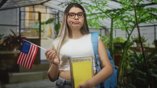 Young woman holds yellow book and usa flag, wearing backpack with pensive mouth expression in greenhouse building; homesick longing.