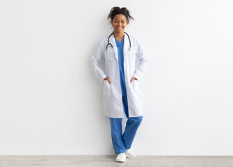 Friendly black woman doctor posing with hands in pockets looking at camera standing leaning on wall, lady wearing blue uniform, coat and stethoscope, full body length, white studio background banner © Prostock-studio