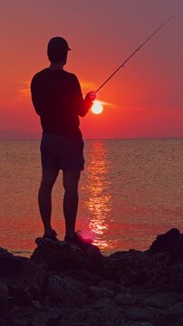 Vertical video. Man Fishing from Rocks Facing Sunset Over Sea Wide Angle Silhouette Outdoor Adventure Coastal Angling Scene