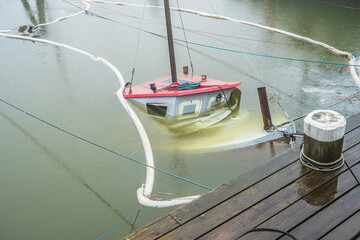 Partially submerged small sailboat surrounded by oil containment boom. © Trygve