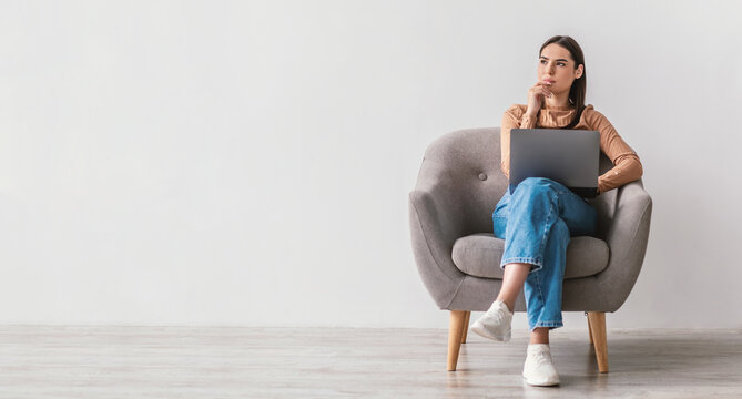 Thoughtful millennial lady with laptop pc sitting in armchair, looking at empty space against white studio wall, banner design. Lovely young woman using portable computer, working online