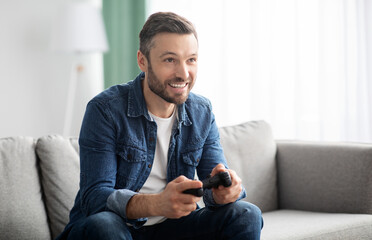 Joyful middle aged bearded man playing video game with joystick at home, sitting on couch in living room, having fun at weekend, copy space. Home entertainment, leisure, domestic lifestyle © Prostock-studio