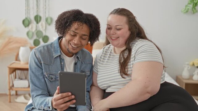Man and plus size woman on bed look at tablet while woman shows hand with ring in a studio; affection bond.