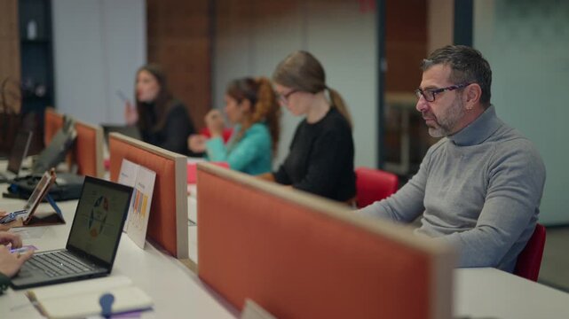 An office environment shows diverse individuals diligently working at their workstations. People are focused on their computers and notebooks in a collaborative workspace.