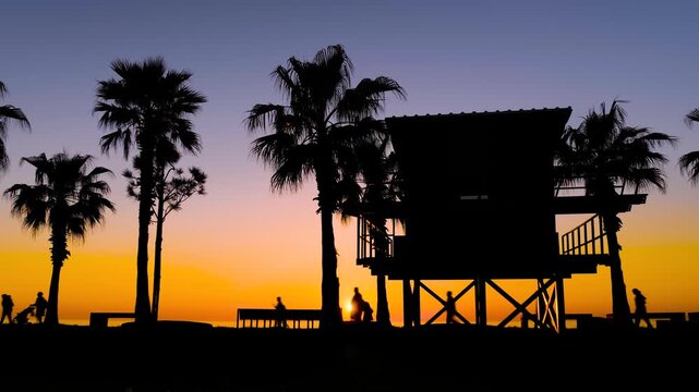 Timelapse: silhouettes of palm trees and lifeguard tower on the embankment of the resort town at sunset. People walking along the waterfront against warm sky - lens flare. Summer and tropical concept
