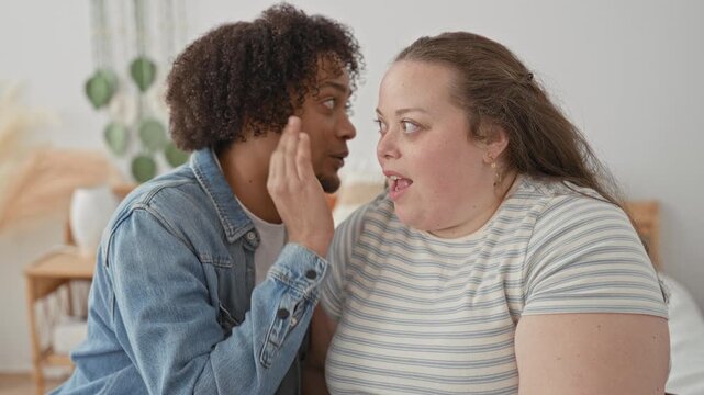 Woman listens as man whispers into her ear, hand near her face on a bedroom bed with plants and nightstand visible; secret affection playful.