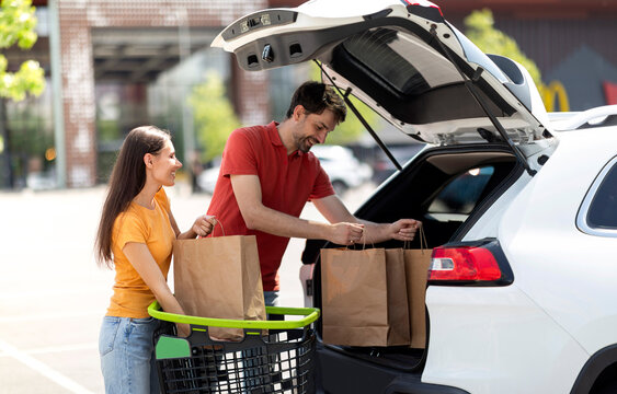 Happy beautiful young caucasian couple buying food in grocery store, packing purchases in white car. Husband and his wife after buying food at supermarket, loading shopping bags in auto trunk