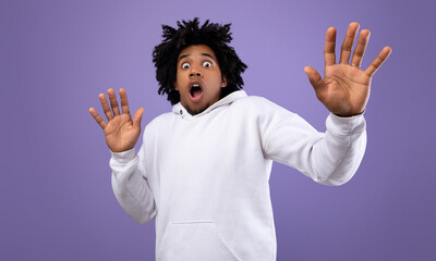 Shocked black guy making protection gesture, feeling frightened on violet studio background,...