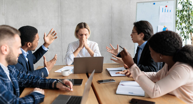 Aggressive Colleagues Shouting At Unhappy Victimized Business Lady Sitting At Desk In Modern Office. Problems And Disagreement At Work, Business Team Communication, Corporate Bullying Issue. Panorama