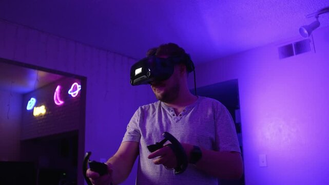 Man in VR headset holding controllers playing virtual reality at home performing movements and interacting with digital environment immersing into gameplay under neon lighting