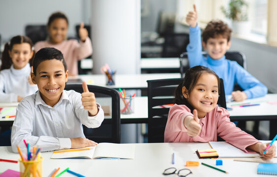 Education Is Fun Concept. Diverse group of happy smiling international classmates sitting at desks in classroom and showing thumbs up sign gesture, children enjoying studying at junior school