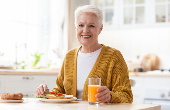 Cheerful attractive grandmother in casual outfit posing in kitchen, having healthy lunch alone, copy space. Happy grey-haired senior woman eating healthy sandwich, drinking fresh juice, home interior