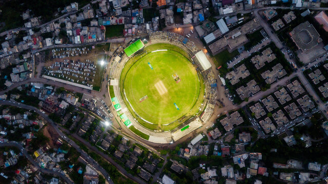 Aerial view of Muzaffarabad Cricket Stadium illuminated by floodlights surrounded by urban buildings and parked cars in Muzaffarabad, Azad Kashmir, Pakistan.