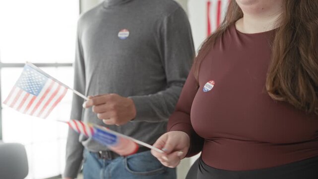 Woman and man holding small american flags and wearing i voted stickers while standing inside a polling station building, hands gripping flag sticks and casual attire visible; civic duty pride.
