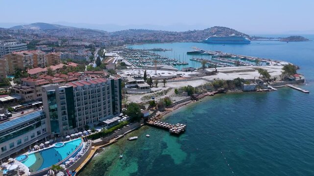Kusadasi Marina in Turkey, showing rows of docked white yachts and sailboats in turquoise water, with a large cruise ship in the background and the coastal city sprawling across the hills.