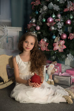 Cute little girl in princess dress is posing at home front of Christmas tree with pomegranate in hand. Christmas and New year concept. Holiday home decoration. Selective focus