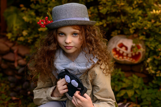 Little cute smiling girl in grey hat with long curly hair holds toy. Autumn seasonal portrait of young child girl outdoor in the garden front of  the autumn Halloween background. Selective focus
