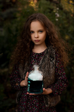 Happy Halloween Concept! Cute little with green witch's potion with smoke in the decorated garden. Beautiful child girl in witch costume and long curly hair. Selective focus, close up