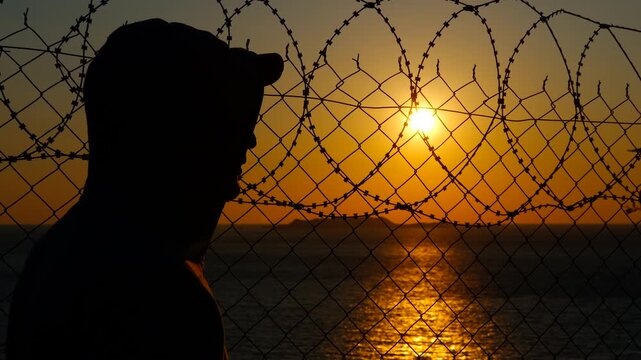 Man observing sunset behind barbed wire fence. Silhouetted refugee standing behind barbed wire fence, gazing across expansive ocean during golden hour, symbolizing hope, and longing for freedom