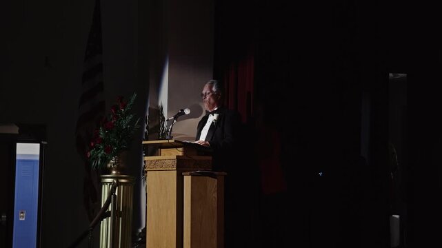 Elderly man delivering a heartfelt speech at a formal event, standing at a wooden lectern on stage