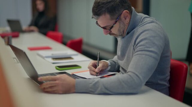 A focused man diligently works at his desk, using a laptop and writing in a notebook. He concentrates on tasks in a modern, collaborative office setting.