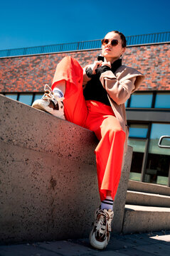 A photograph of a fashionable young woman in a beige jacket, black shirt, and red pants, posing confidently on a sunlit city street. Contemporary urban style and street fashion.