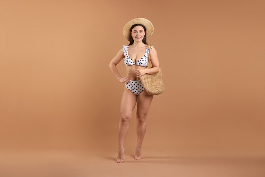 Beautiful woman wearing stylish swimsuit, hat and bag on brown background