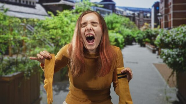 Woman holds yellow socks away from face on a city street planter walkway, grimacing with mouth open and closed eyes; disgust.