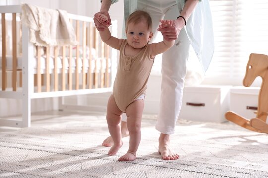 Baby's first steps. Mother supporting daughter while she learning to walk at home, closeup