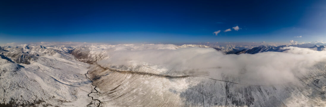 Aerial view of Babusar Pass winding through snow-capped mountain peaks under a clear blue sky with rolling clouds in Babusar, KPK, Pakistan.