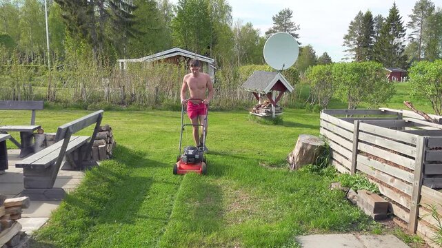 Muscular young man without a shirt pushing a gasoline lawn mower to cut the green grass in his backyard during a bright and sunny summer day, engaging in essential yard maintenance