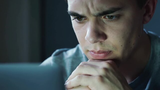Young man looking at computer screen with focused expression in dark room, close up shot