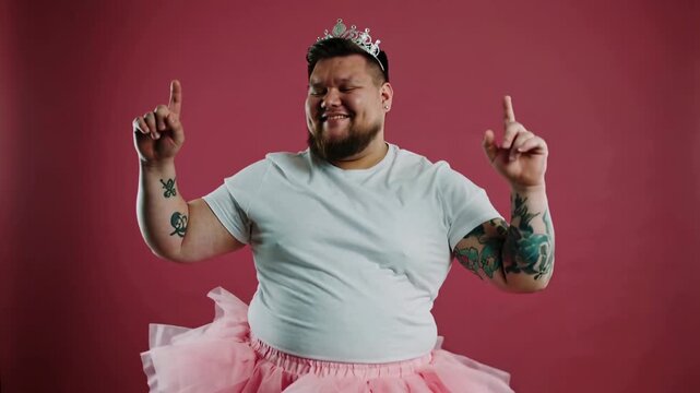 Man in white t-shirt and pink tutu wearing tiara dancing against dark red background, medium shot