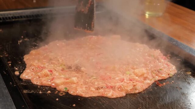 Close-up of a chef's hands using metal spatulas to cook authentic Tokyo-style Monjayaki on a sizzling teppan griddle. The footage captures the steam, the gooey texture of the savory pancake batter, an