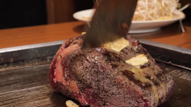 close-up shot of a professional Japanese chef preparing a premium marbled beef steak on a hot iron teppanyaki grill. The chef is seen basting the juicy meat with a generous pat of melting butter and a