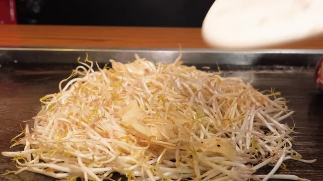 A close-up shot of fresh, crisp mung bean sprouts being skillfully tossed and stir-fried with a metal spatula on a hot commercial griddle. This footage highlights traditional Asian street food prepara