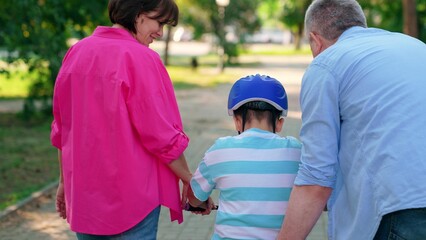Naklejka premium Child son cyclist with parents in city park. Happy family, child in helmet, mother father playing with bicycle outdoors in summer. Family day out. Dad mom teach son to ride bike in park. People park