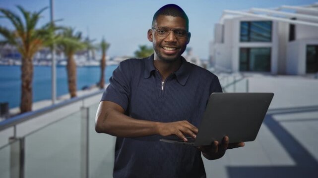 Man typing on laptop while holding the computer and smiling on a building terrace with palm trees and marina visible; confidence.