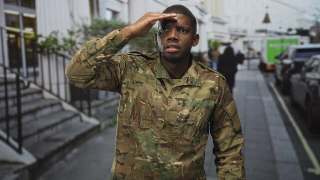 Man soldier in camouflage uniform with hand to forehead shading eyes on a city street beside parked cars and building steps, looking ahead; duty vigilance.