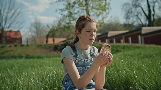 Young girl with freckles sitting in a grassy field and eating a delicious ice cream sandwich