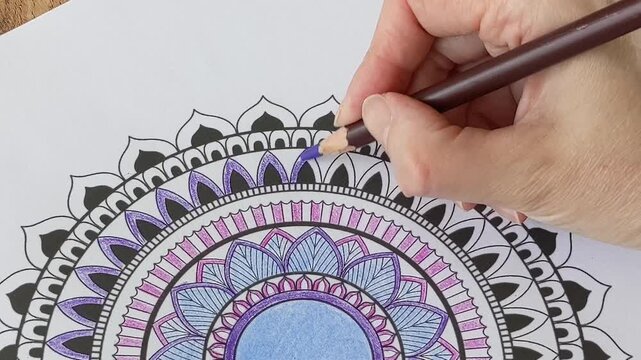 Hand carefully coloring an intricate mandala pattern with a purple colored pencil. Concept of creativity, meditation, art therapy, and stress relief through coloring.