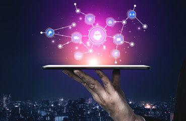 Obraz premium A hand holds a tablet displaying digital icons and connections against a night cityscape. This image represents technology, innovation, and modern communication concepts. Tessel