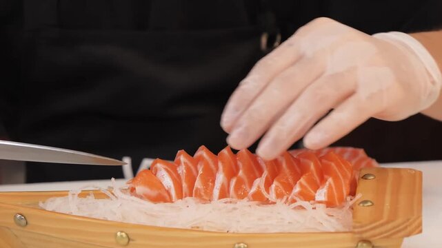 A close-up shot of a professional sushi chef's hands, wearing gloves, meticulously arranging thin slices of vibrant, raw orange salmon sashimi on a bed of shredded white radish. The fresh seafood is p