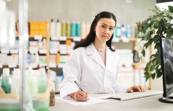 Attentive middle-aged pharmacist making notes on paper for restocking in chemist's shop