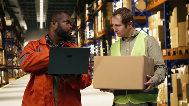 Coworkers arranging cardboard boxes on shelves and doing inventory on laptop, distribution center efficiency depends on workforce collaboration and safety. Logistics management systems.