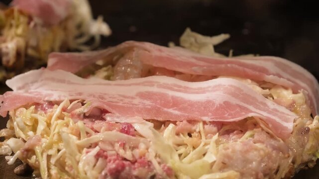 A close-up shot capturing the authentic process of making Hiroshima-style okonomiyaki. The footage shows raw bacon strips layered over a mountain of shredded cabbage and ingredients on a hot metal tep