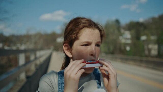 Young girl with freckles wearing overalls playing a harmonica while walking across a bridge in summer